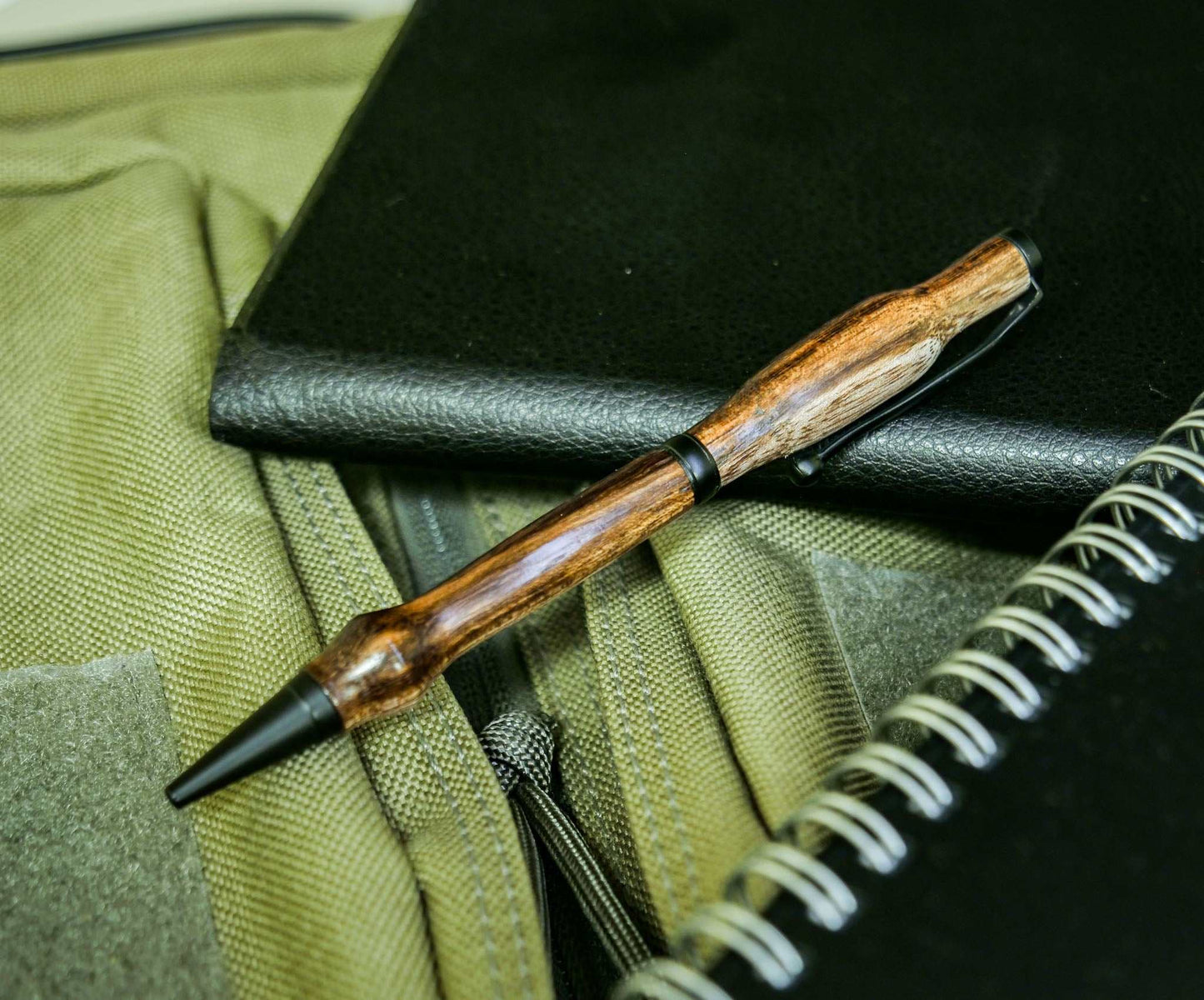 Handmade twist pen with unknown wood body and black enamel hardware placed on green fabric and notebooks