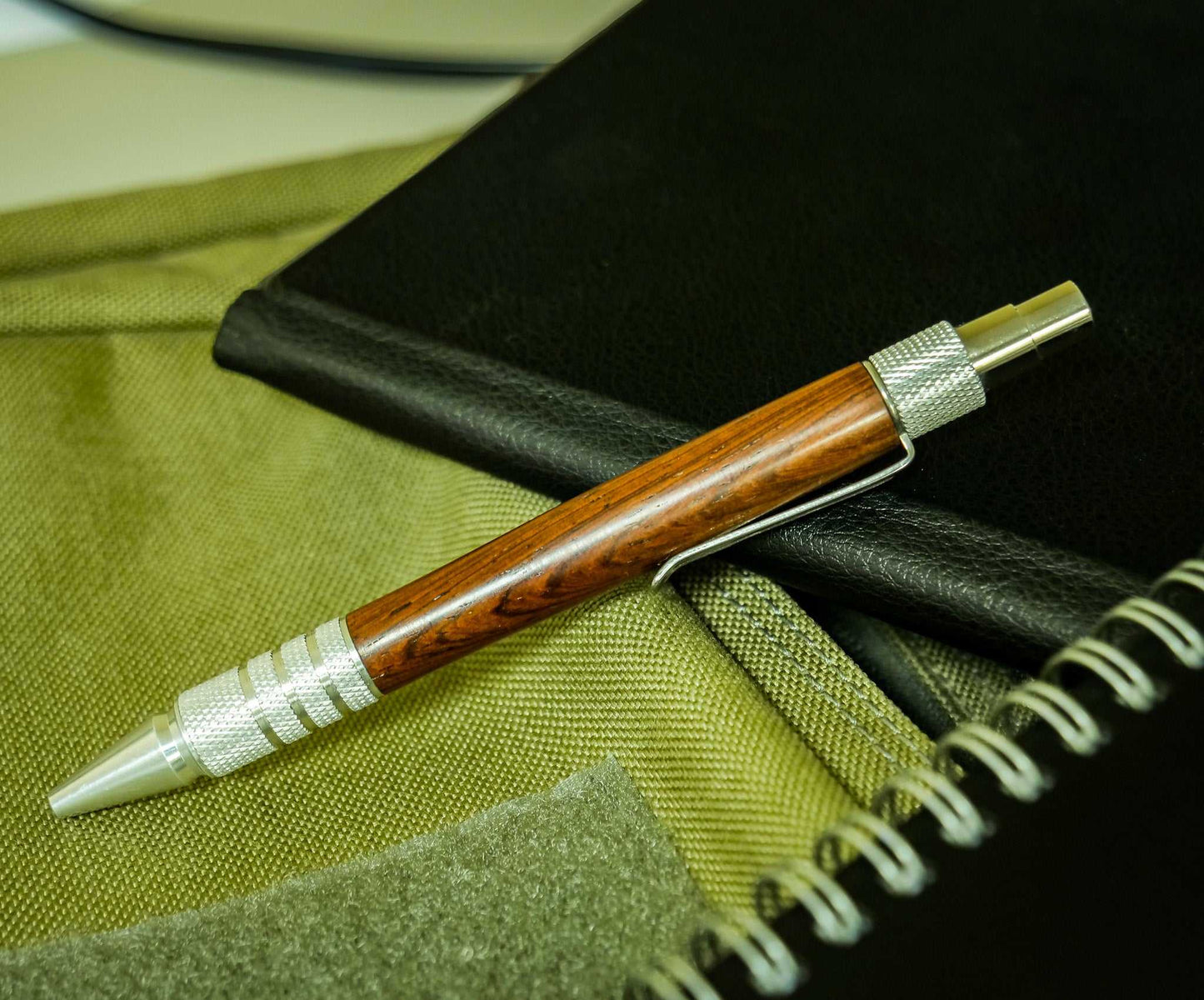 Handmade pen by Ryan Wilson with rosewood body, aluminum hardware, and DuraClick mechanism lying on notebooks.
